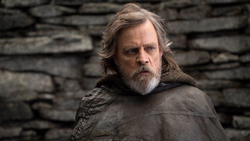 Mark Hamill as an older Luke Skywalker with gray hair and beard, wearing weathered robes in front of a stone wall.