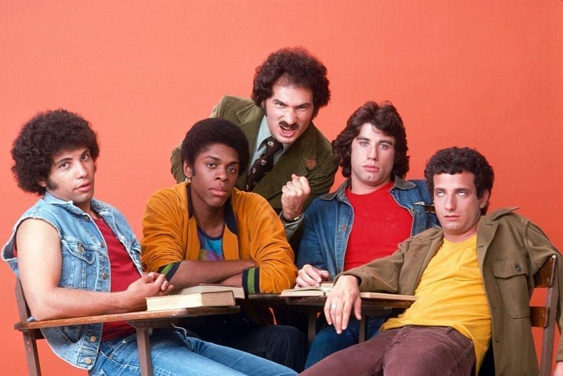 Cast of Welcome Back, Kotter, with Mr. Kotter posing behind four students seated at school desks against an orange backdrop.