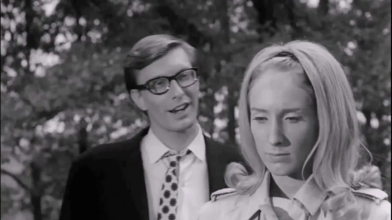 Black-and-white still from Night of the Living Dead: a grinning man in glasses leans in behind a somber blonde woman in a graveyard.