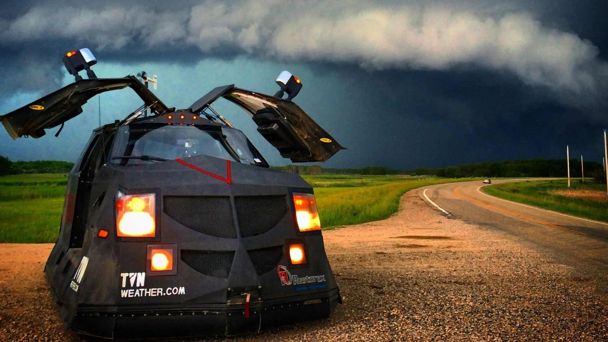 A black armored storm-chasing vehicle with gull-wing doors raised, parked on a gravel shoulder while a dark storm shelf hangs over the road behind it.